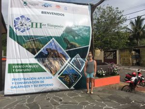 Gillian Munro stands in the street next to a giant banner announcing the "IIIer Simposio" in Galapagos. 