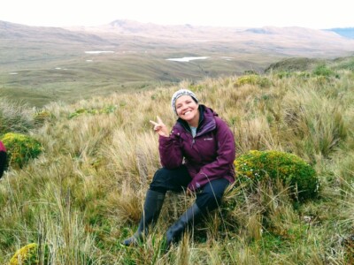 Photo: A smiling woman wearing a purple jacket and black wading boots flashes a 'peace' sign in the foreground of a mountainous landscape.