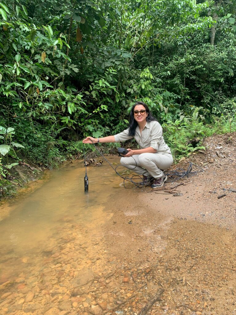 A woman in head-to-toe khaki dangles a sampling device from a long black cord into a cloudy stream.