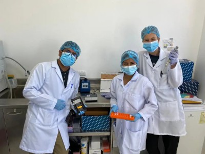 Three researchers in full PPE equipment stand in a lab, holding (l-r) a small black bag, an orange tray, and a pipette.