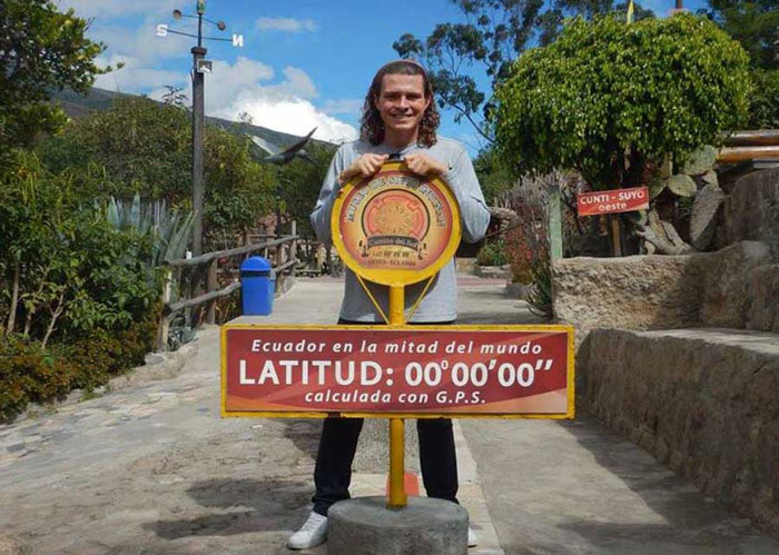 Ryan Schmedding resting his hands on a sign reading 'Ecuador in la mitad del mundo' on a town's dirt road.