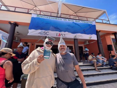 Two people wearing shark hats pose in front of a Shark Week banner.