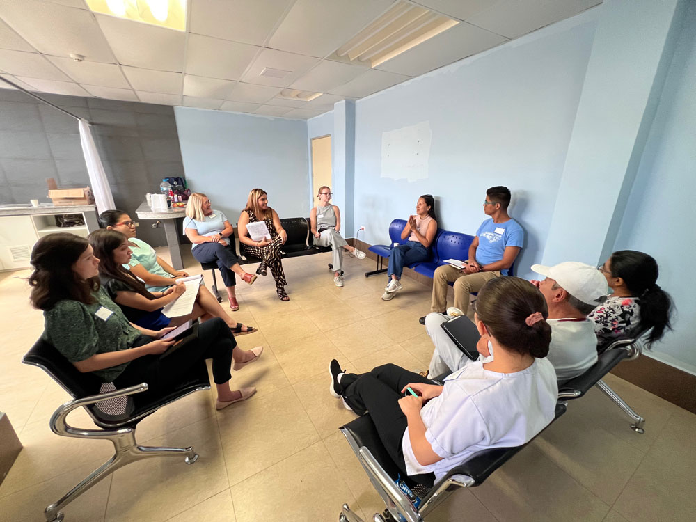 A group of 11 people sit in a square cluster of chairs, having a discussion. 