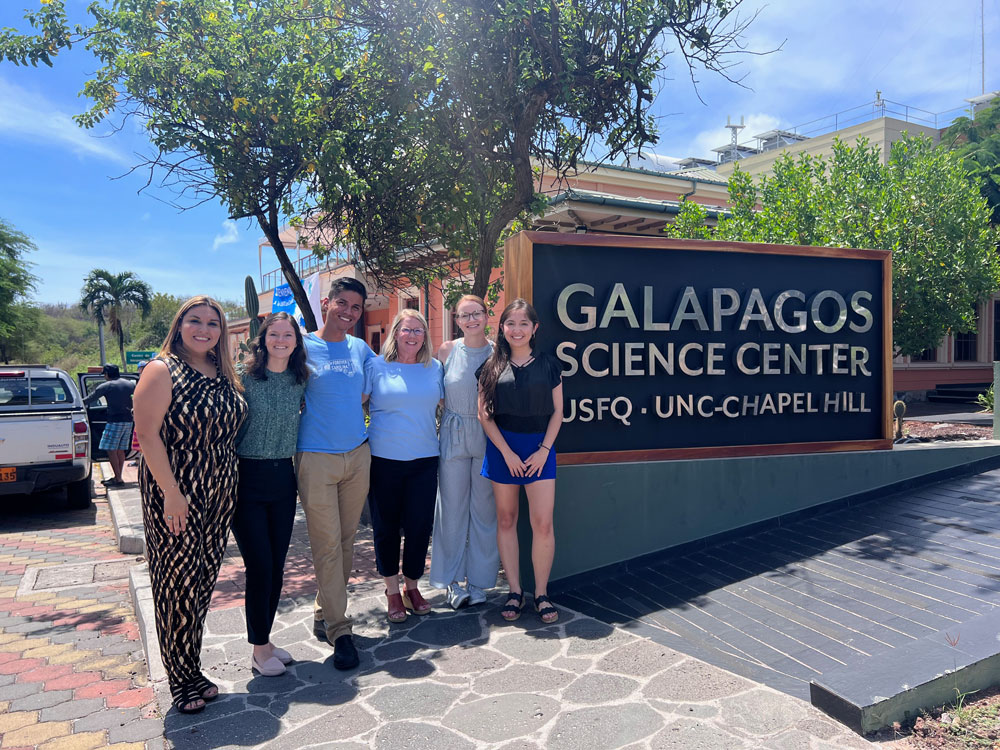 6 students and faculty researchers stand next to a sign reading "Galapagos Science Center" and "USFQ | UNC-Chapel Hill" outside of the center.