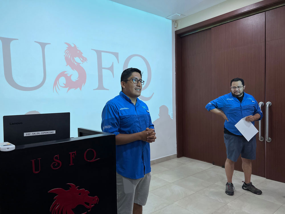Andrés Pazmiño lectures in front of a projector screen displaying the USFQ school logo. Carlos Mena watches from the right.