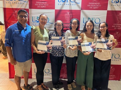 Andrés Pazmiño (left) poses with five program participants, who are holding up their program certificates, in front of a USFQ-branded banner.