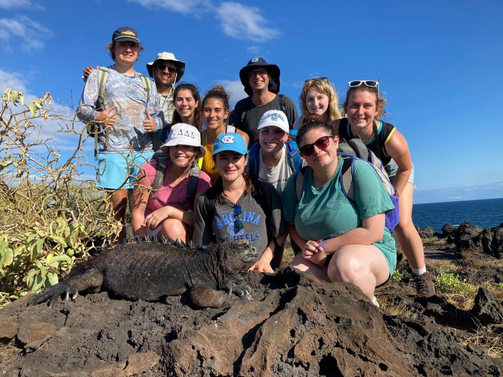 A group of 11 students pose in the Galapagos landscape, a native iguana posing on a rock in front of them.