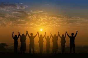 Stock photo of a line of teenager figures, details not visible in the sunset, holding hands and raising their arms in the sky. 