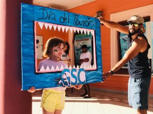 A young child wearing face paint stands behind a blue frame with paper "teeth" and the words "Dia del Tiburon" (shark day).