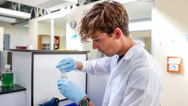 A graduate student researcher, wearing a lab coat and gloves, handles a test sample in the lab.